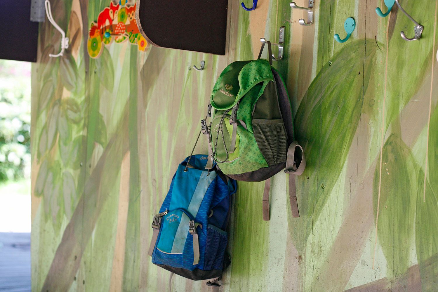 Naturkita Waldforscher - Auf der mit Blättern bemalten Holzwand der Schutzhütte hängen ein grüner und ein blauer Kinderrucksack für den Wanderausflug.