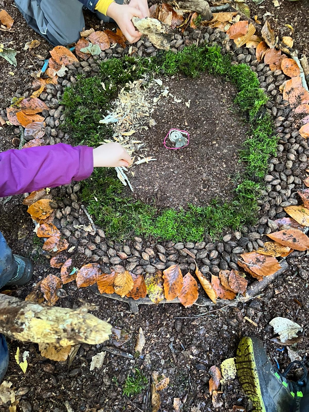 Naturkita Waldforscher - Kinder legen ein Mandala aus Eicheln, Moos, Blaetern, Steinen und Bucheckern auf den Waldboden.
