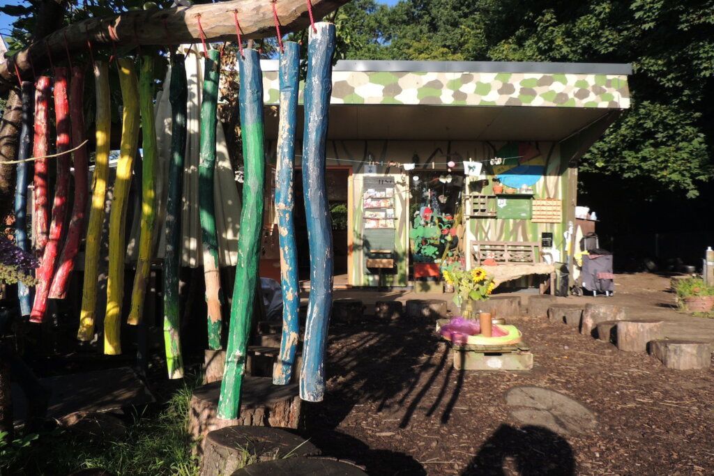 Waldforscher Naturkita - Vor der Schutzhuette im Sonnelicht hängt eine bunte Stoeckchengirlande in den Regenbogenfarben.