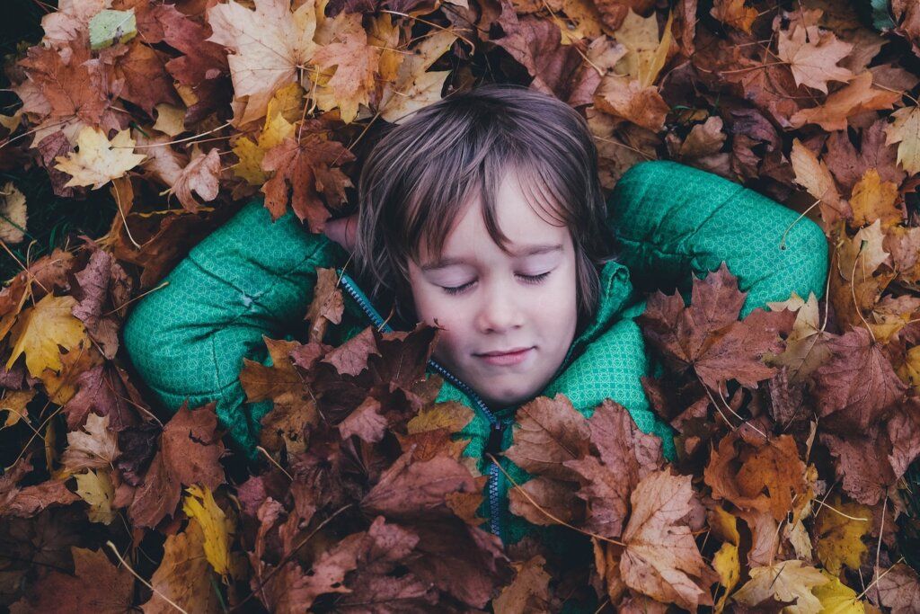 WALDFORSCHER Waldkindergarten - Ein Kind liegt mit geschlossenen Augen und mit hinter dem Kopf verschränkten Händen im Herbstlaub.