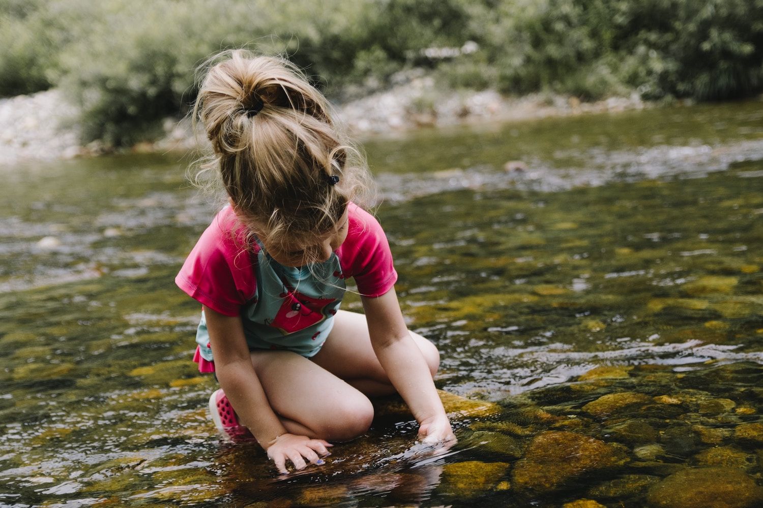 WALDFORSCHER Waldkindergarten - Ein Mädchen in Sommerkleidung und mit Pferdeschwanz spielt in einem seichten Bach. Nachhaltiges Erleben in unserer Naturkita.